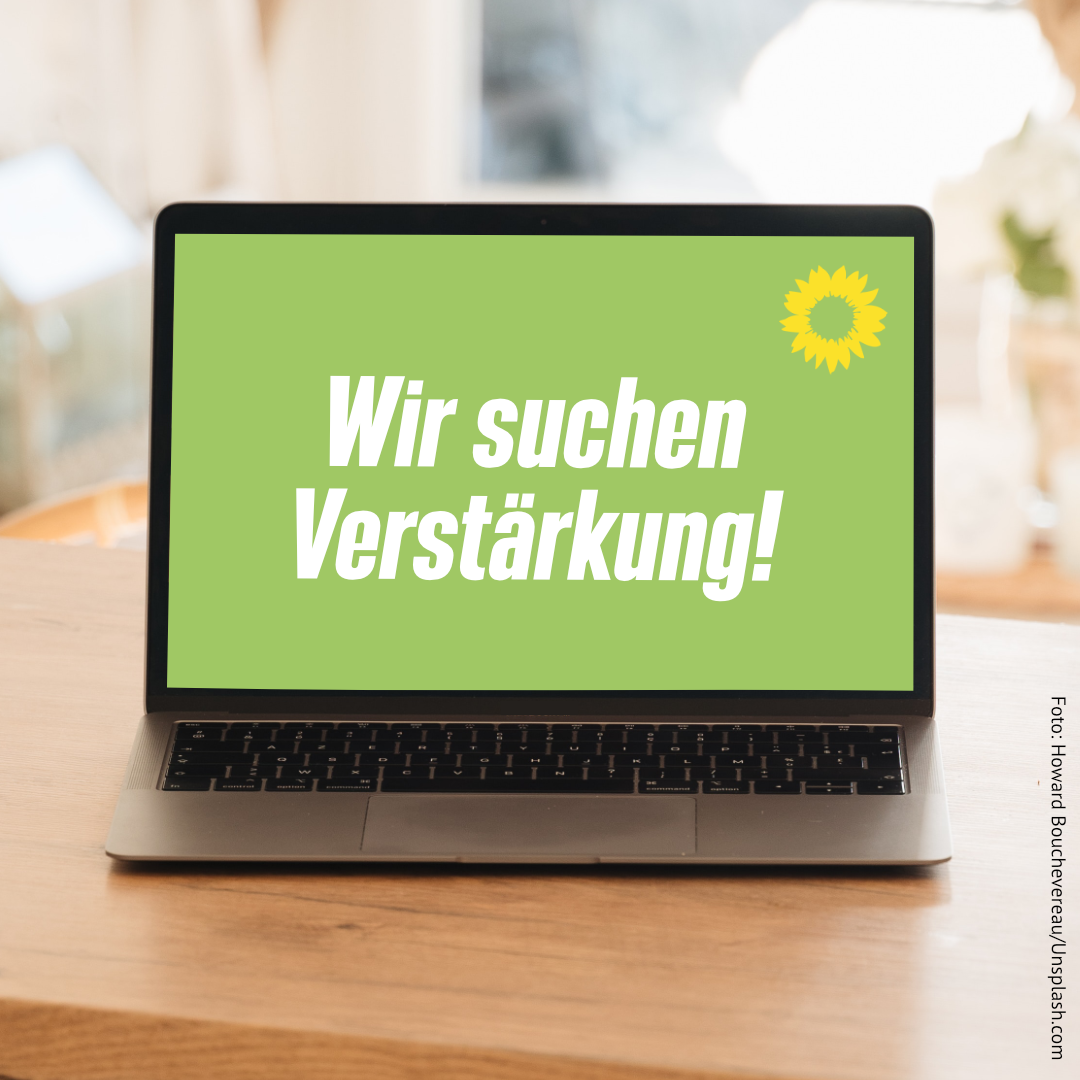 Ein Laptop steht auf einem Holztisch. Auf dem Bildschirm befindet sich ein grünes Bild mit der Aufschrift "Verstärkung gesucht". Darüber ist eine Sonnenblume.