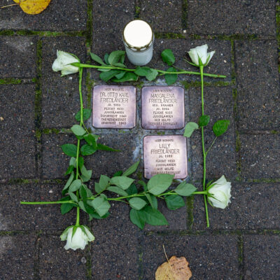 Die Stolpersteine in der Bahnhofstraße werden von weißen Rosen eingerahmt. Daneben steht eine Kerze.
