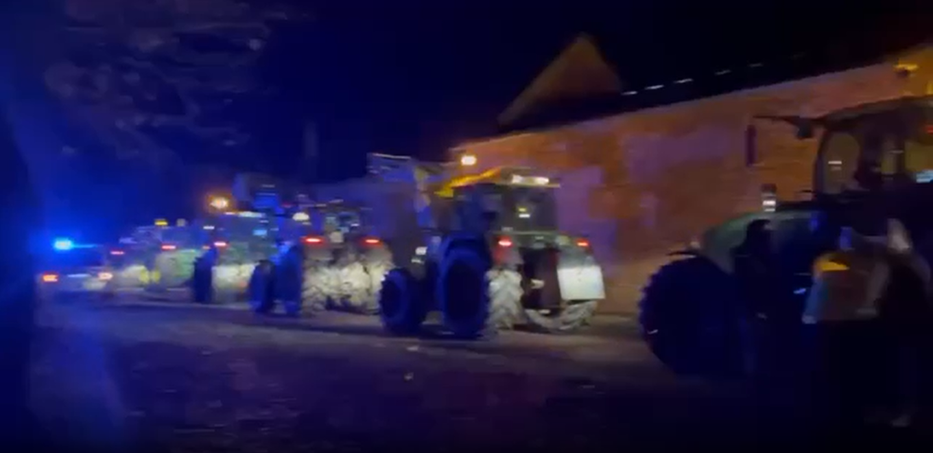 Traktoren und ein Polizeiauto stehen im Dunkeln mit eingeschalteten Lichetern auf einer Straße.