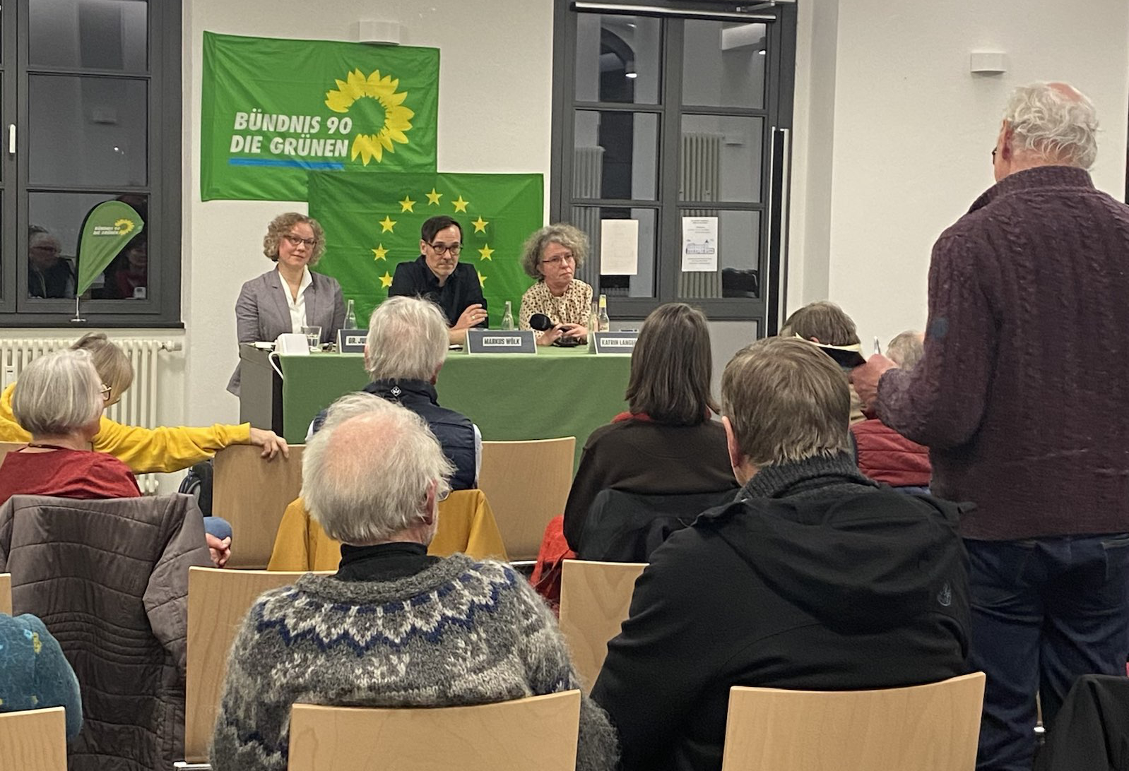 3 Personen sitzen am Podiumstisch, davor sitz das Publikum. Ein Mann steht und redet. Im Hintergrund die Flagge der Bündnis 90 die Grünen