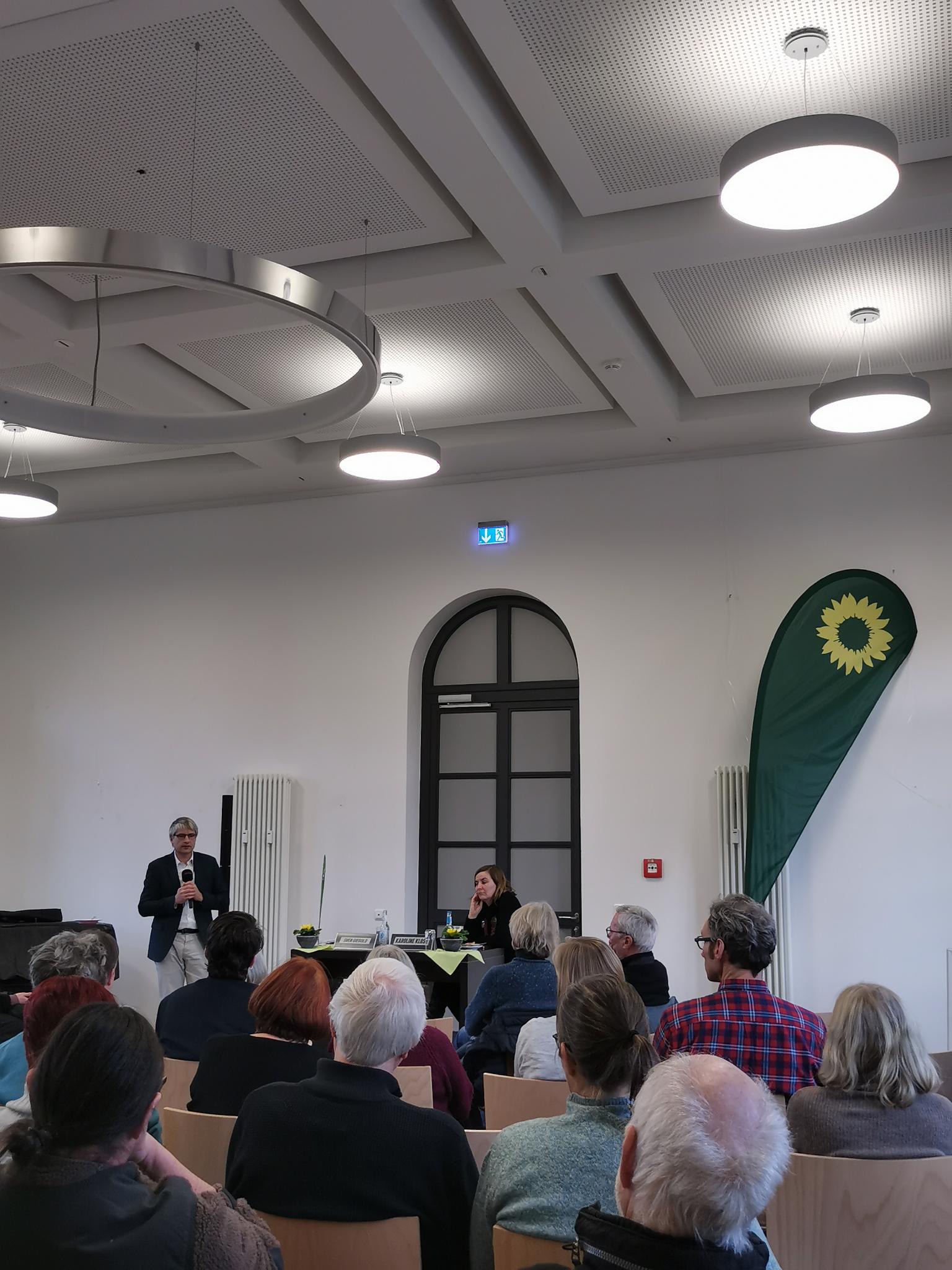 Sven Giegold steht im Hintergrund und hält eine Rede. Rechts steht eine große grüne Fahne. Davor sitzen Menschen mit dem Rücken zum Fotografen.